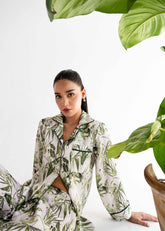 A woman sitting with a tropical leafy patterned sleepwear set, consisting of a button-up shirt and matching pants, with a large green leaf in the background.